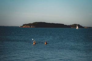 Two people kayaking in Stockholm archipelago with a sailboat and islands in the background.